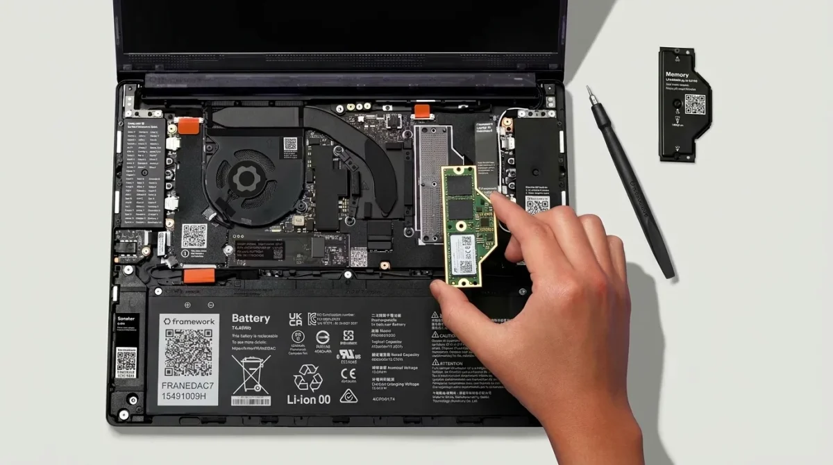 An overhead view showing the internal layout of the open Framework Laptop 13 Pro (2026). A hand holds the swappable LPCAMM2 memory module over the mainboard. Visible components include the new massive 74Wh battery, the updated dual-fan cooling system for Panther Lake processors, and a QR code linking to the interactive repair guide.
