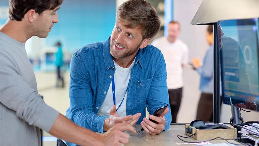 A Virgin Media O2 store representative in a blue denim shirt helping a customer with a mobile phone at a tech support desk.