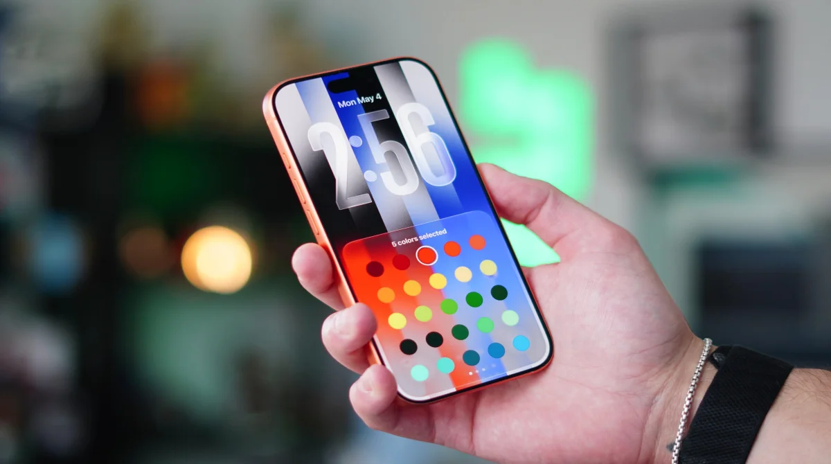 A hand holding an iPhone displaying the new Pride Luminance wallpaper and a dynamic color selection palette, highlighting the UI personalization features available in Apple iOS 26.5.
