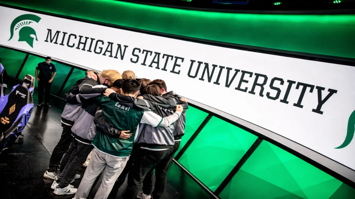 The Michigan State University esports team huddles in a circle on stage in front of a giant green and white screen reading Michigan State University, showcasing a competitive match day for msu gaming.