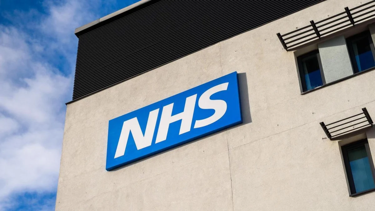 A large blue and white NHS sign on the exterior of a modern medical facility, representing the physical infrastructure supported by the NHS App.