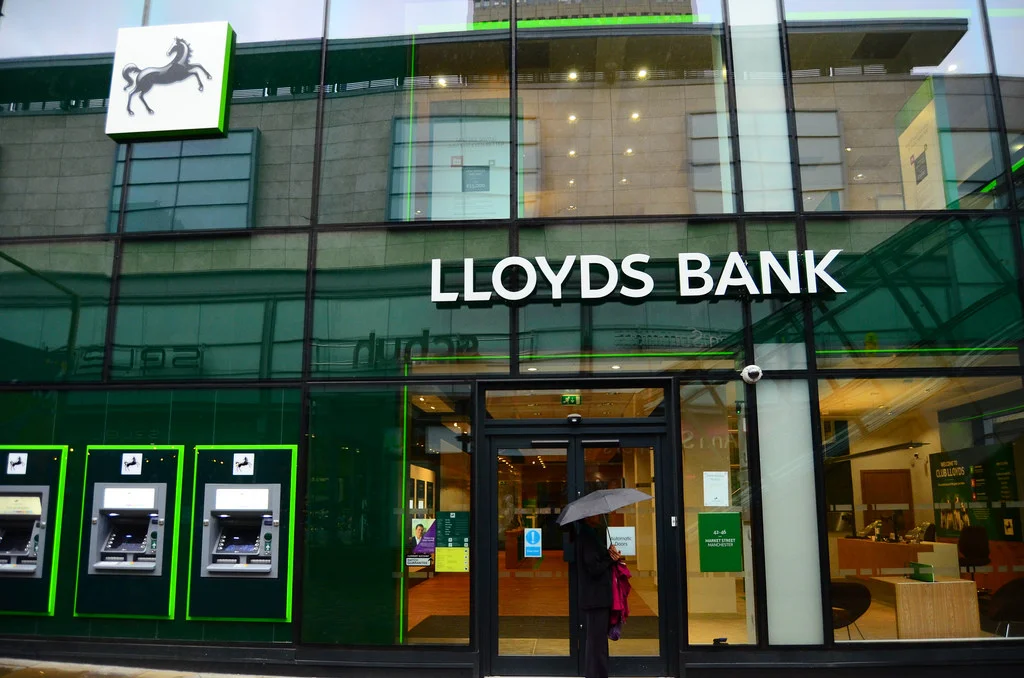 Exterior of a glass-fronted Lloyds Bank branch on Market Street Manchester with bright green panels, cash machines, and a customer holding an umbrella, used to highlight Lloyds Bank issues.